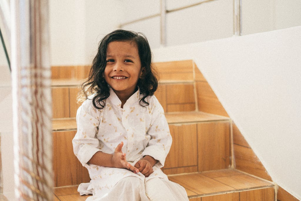 Charming child with long black hair sitting and smiling on wooden stairs indoors.